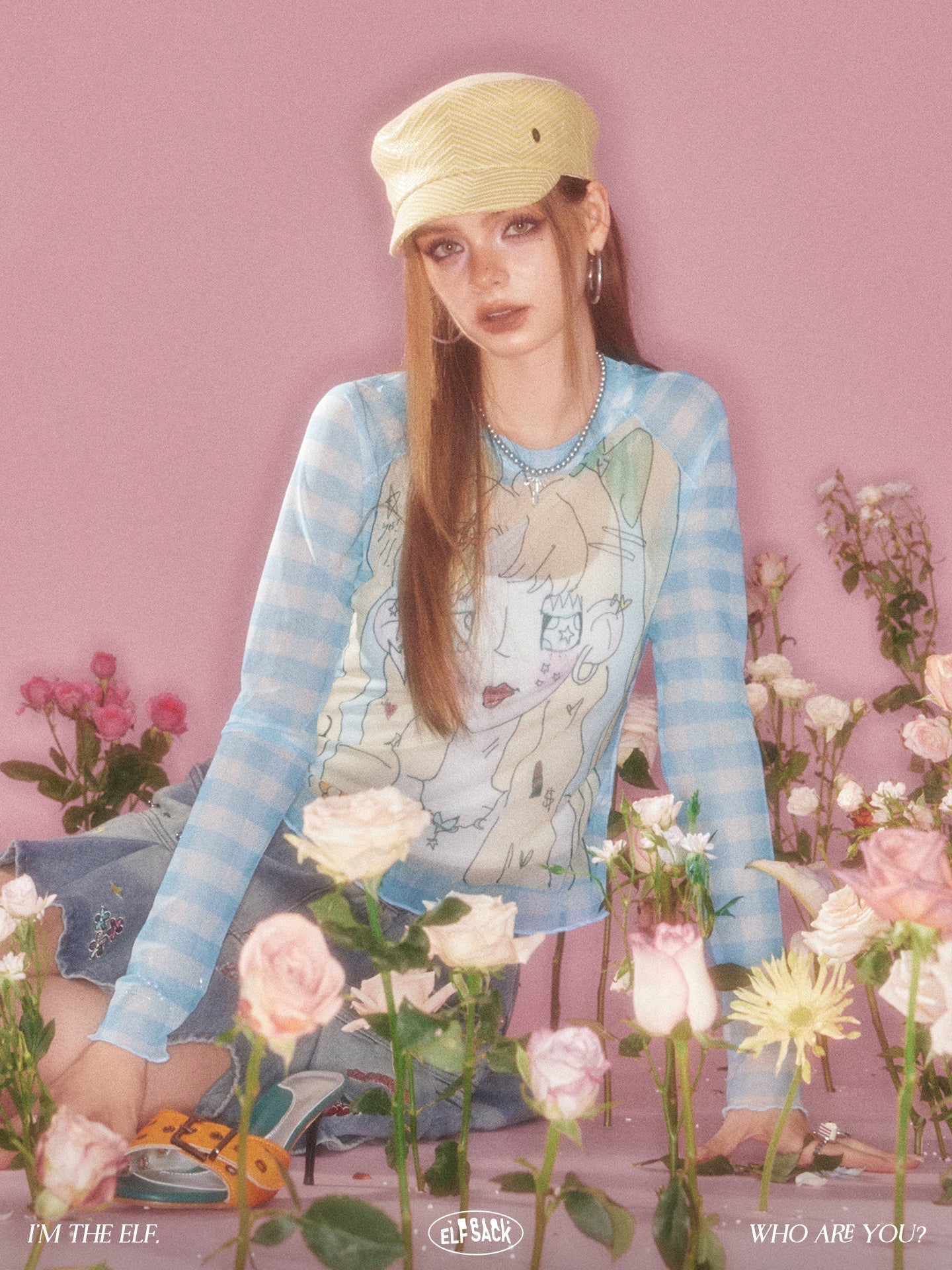 Young woman wearing a pastel blue graphic sweater and yellow cap surrounded by pink and white flowers